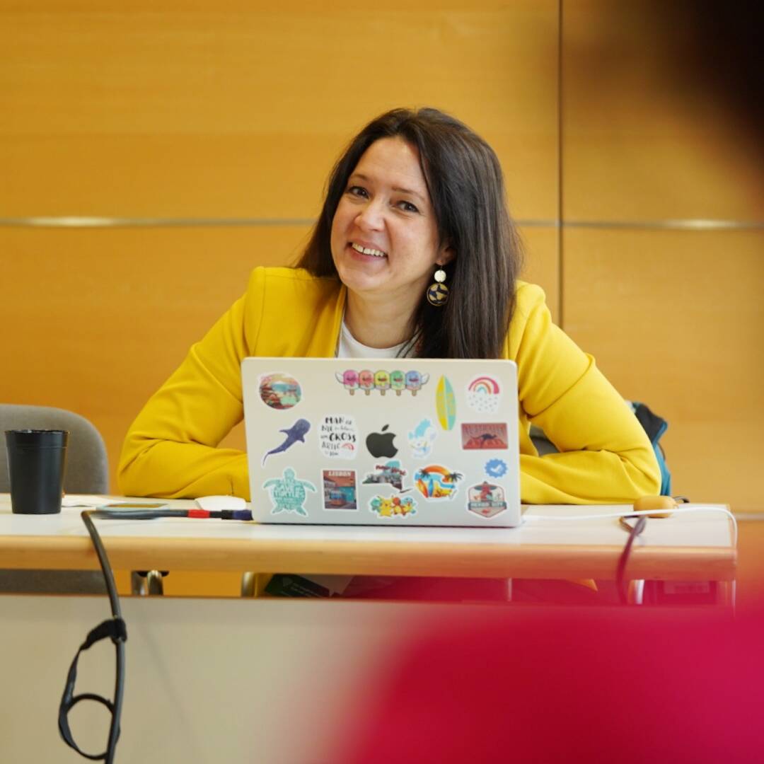 Daniela Vey hält einen Workshop. Vor ihr auf dem Tisch steht ihr MacBook. Sie trägt ein gelbes Jacket und lacht mit den Teilnehmerinnen.
